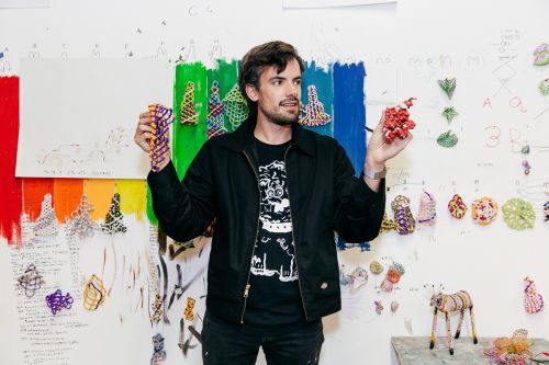Adult male holding colorful beaded art in an artist studio with drawing covered walls, wearing a black jacket and patterned t-shirt.