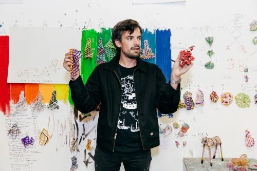 Adult male holding colorful beaded art in an artist studio with drawing covered walls, wearing a black jacket and patterned t-shirt.
