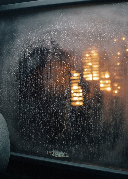 Close-up of a rain-streaked window pane with blurred warm city lights glowing in the dark background.