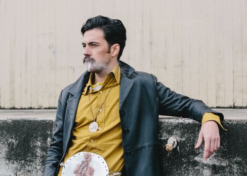 Adult male smoking cigarette outdoors, wearing a black leather coat and yellow shirt against a concrete wall in medium shot.