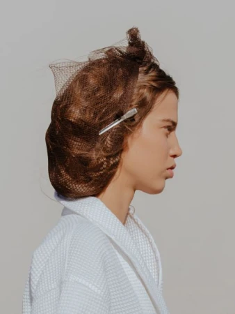 Profile view of a light-skinned young adult woman with brown hair wearing a white bathrobe, indoors, with a neutral expression.