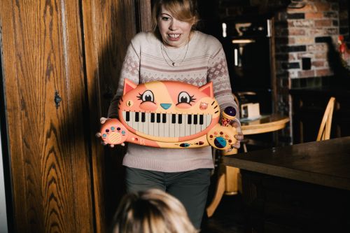 Young woman holds a cat-shaped toy piano indoors. A child is partially visible in the foreground. The background includes a wood door and brick wall.