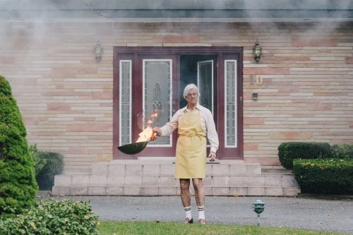 Elderly man wearing a yellow apron holds a frying pan with open flames in front of a brick house with smoke drifting.