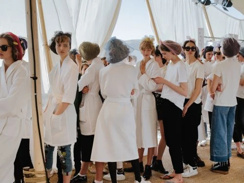 Group of young women in white robes and hairnets backstage at a fashion show, standing near white curtains, getting ready for the show.