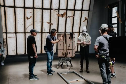Clown in costume surrounded by film crew on set with prop moths in front of window during filming in a studio environment.