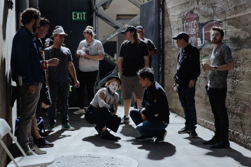 Group of people on film set, including performer in makeup. Actors, film crew, and equipment visible in industrial-style setting.