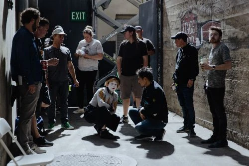 Group of people on film set, including performer in makeup. Actors, film crew, and equipment visible in industrial-style setting.