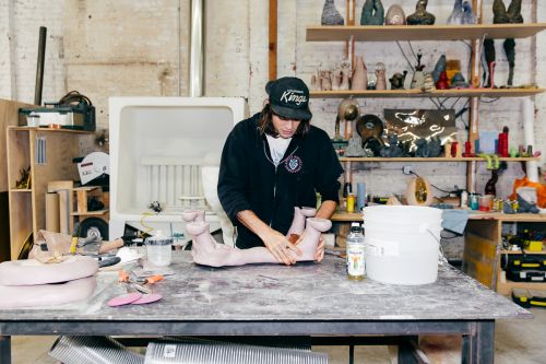 Sculptor shaping pink clay form in a bright workshop; shelves filled with art pieces line the background, bathed in natural light.