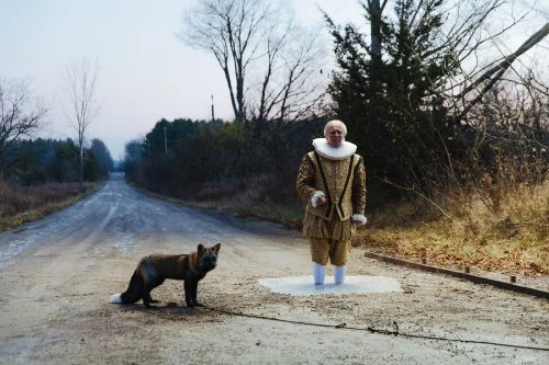 An adult male in period clothing stands on a dirt road with a fox in front of a tree line under an overcast sky.