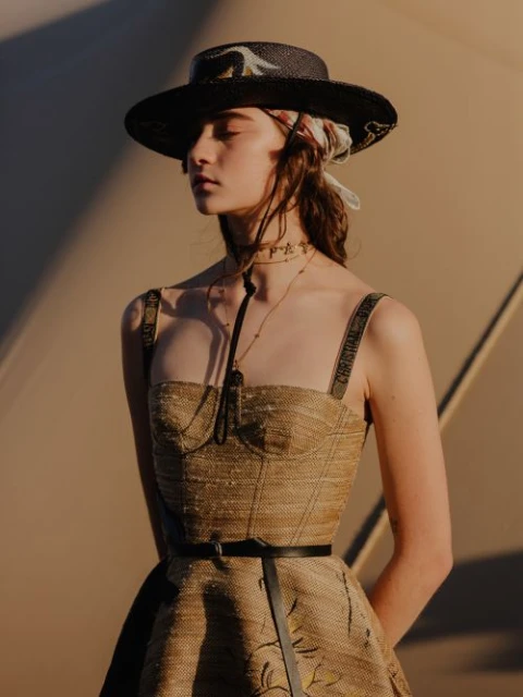Woman poses in a straw-textured dress with Dior straps and a straw hat, eyes closed, against a tan studio backdrop with subtle lighting.