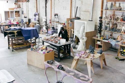 Indoor view of an artist working on sculptures in a bright studio. There are numerous art pieces, tables, and equipment in the workspace.
