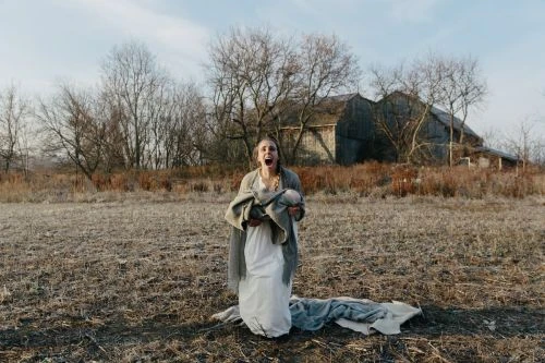 A distressed woman in a white dress stands in a field, holding a baby in a blanket, with an old barn and trees in the background.