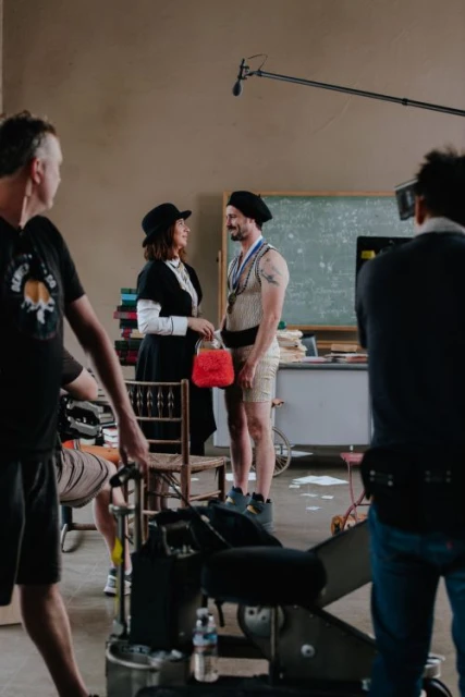 A film set scene with a woman in vintage attire holding a red purse, and a man in a vintage athletic outfit, on film set with crew.