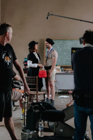 A film set scene with a woman in vintage attire holding a red purse, and a man in a vintage athletic outfit, on film set with crew.