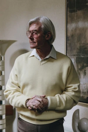 Elderly man with silver hair wearing glasses and a pale yellow V-neck sweater standing indoors