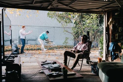 Man in chair playing video game in driveway; two men in background, TV, sofa, coffee table, brick wall, and leafy trees.
