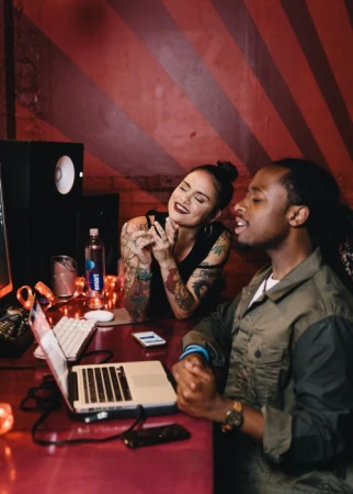 Two people in a music studio: a woman with tattoos smiles as a man sings into a microphone near a laptop, sound equipment, and striped wall.