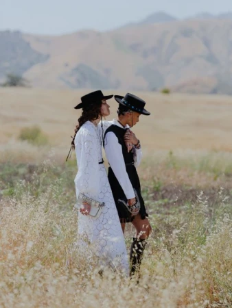Two women wearing wide-brimmed black hats and dresses standing in a dry grassy meadow with distant hills under a pale sky.
