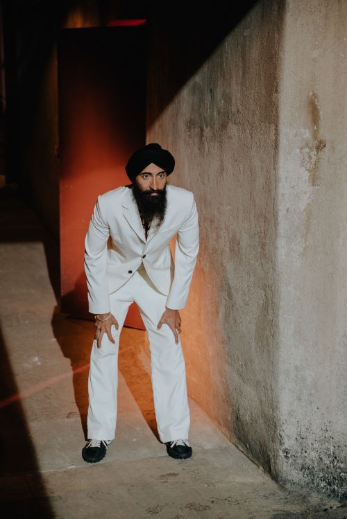Adult man in a black turban and white suit poses in an indoor corner with neutral walls, one knee bent, in an eye-level medium shot.