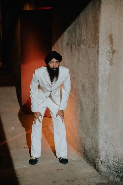 Adult man in a black turban and white suit poses in an indoor corner with neutral walls, one knee bent, in an eye-level medium shot.