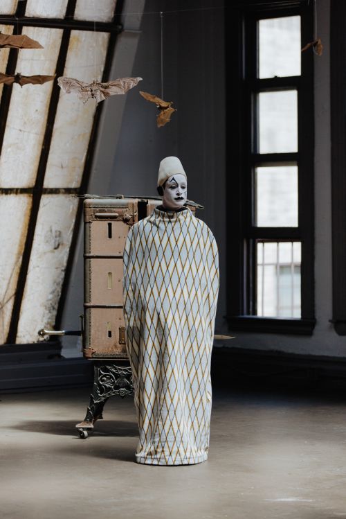Person in clown costume stands in room with diamond pattern robe, white cap, makeup, and bats hanging in front of window.