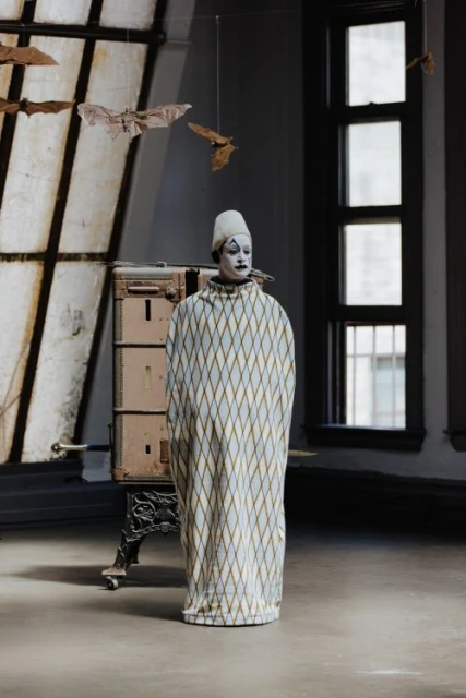 Person in clown costume stands in room with diamond pattern robe, white cap, makeup, and bats hanging in front of window.