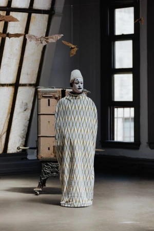 Person in clown costume stands in room with diamond pattern robe, white cap, makeup, and bats hanging in front of window.