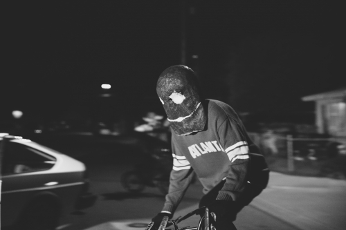 Monochrome shot of a person in a mask riding a bicycle at night, featuring urban environment and a vintage car in the background.