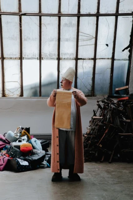 An adult stands indoors holding a tan paper roll. The person wears a long pink sweater and white cap in front of a window and objects.