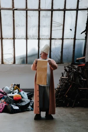 An adult stands indoors holding a tan paper roll. The person wears a long pink sweater and white cap in front of a window and objects.