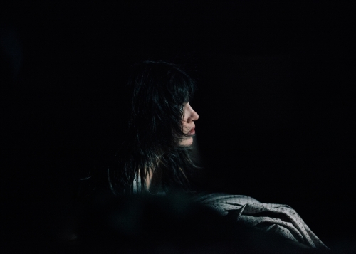 Profile portrait of a woman with dark hair in a dimly lit, dark environment. The woman is facing left with soft lighting.