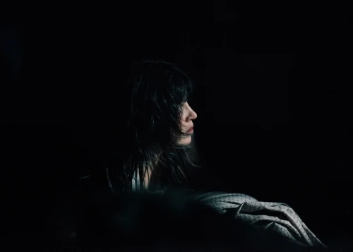 Profile portrait of a woman with dark hair in a dimly lit, dark environment. The woman is facing left with soft lighting.