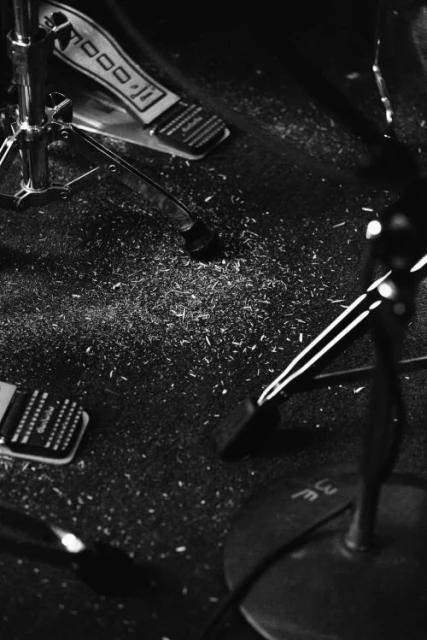Monochrome shot of a drum set with a pedal, cymbal stand, and drumsticks on a dark surface with scattered debris.
