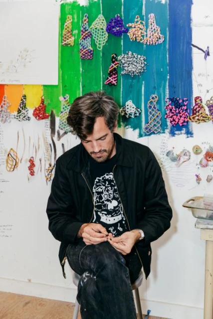 Adult male artist with beard sits on stool in art studio, working with wire. Colorful art and sketches on white walls.