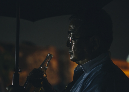 Silhouette of an adult male holding a bottle under an umbrella at night, creating a moody and contemplative outdoor scene.