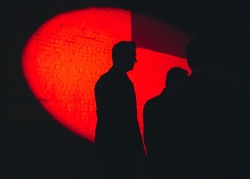 Silhouetted figures stand against a red spotlight on a textured wall, creating a dramatic and high-contrast visual.