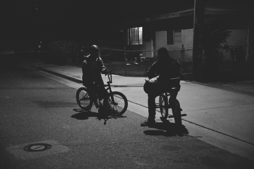 Two figures on BMX bikes pause on a dark street at night, illuminated by a nearby window, creating a monochrome scene with long shadows.