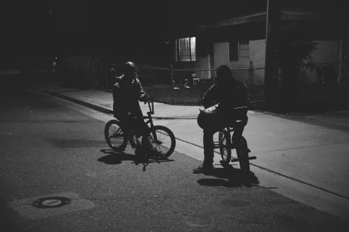 Two figures on BMX bikes pause on a dark street at night, illuminated by a nearby window, creating a monochrome scene with long shadows.
