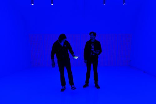 Two adults stand in a room illuminated with blue light. The room features blue walls, and the people are holding a cell phone.