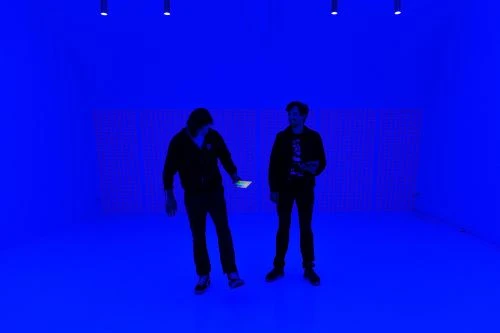 Two adults stand in a room illuminated with blue light. The room features blue walls, and the people are holding a cell phone.