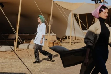 Two models walking outside large canvas tents wearing head coverings and coats during a fashion backstage scene
