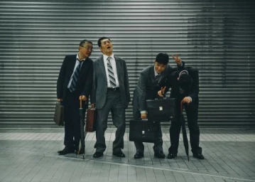 Four businessmen in suits holding briefcases posing playfully in front of a closed metal shutter door.