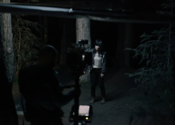 Behind-the-scenes shot of a filmmaker operating a camera rig in a dark forest at night facing a woman standing among tall trees