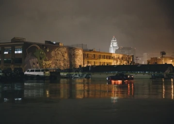 Nighttime street scene of a wet parking lot reflecting city lights, with a car, murals on a building, and a distant illuminated tower.