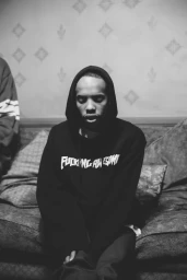 Black and white portrait of a man in a black hoodie seated on a patterned sofa, captured in Los Angeles by Joyce Kim.