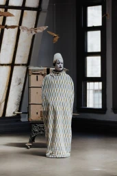 Mime in white face paint wearing a diamond-patterned cocoon garment in a studio space with suspended paper bats.