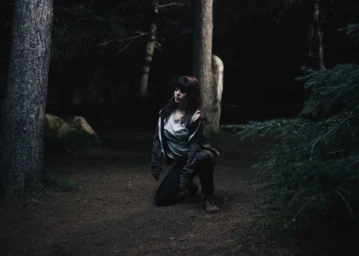 A woman crouching on a dark forest path surrounded by tall trees and pine branches under dim lighting.