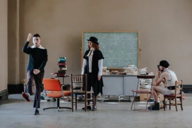 Three performers in theatrical costumes pose inside a large room with a chalkboard, books, and scattered chairs.