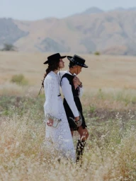 Two women wearing wide-brimmed black hats and dresses standing in a dry grassy meadow with distant hills under a pale sky.