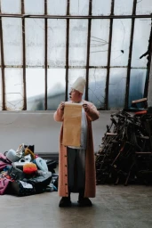 A person in a tall white hat and pink coat holding up a long paper scroll in an industrial space.